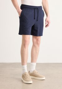 Pantalones cortos azul marino con cintura elástica y cordón; bolsillos laterales; textura suave. Llevados con zapatillas beige y calcetines blancos de canalé.