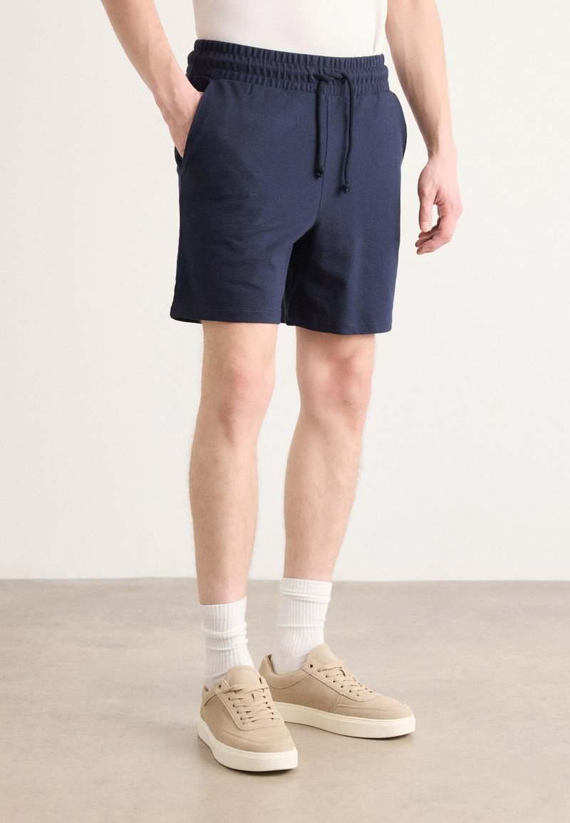 Pantalones cortos azul marino con cintura elástica y cordón; bolsillos laterales; textura suave. Llevados con zapatillas beige y calcetines blancos de canalé.