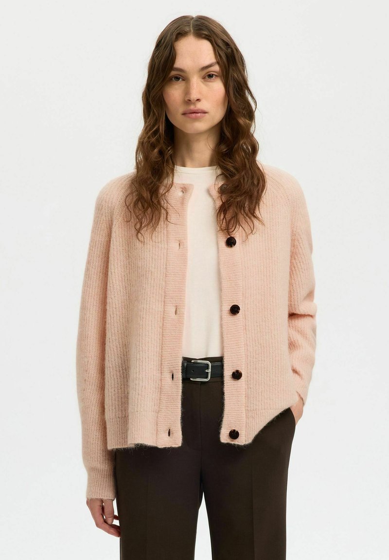 Femme aux longs cheveux ondulés portant un cardigan boutonné rose pâle sur un haut blanc et un pantalon noir avec une ceinture noire, debout devant un fond uni.