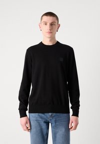 Zwart gebreide trui met een ronde hals, lange mouwen en geribbelde manchetten. Bevat een klein vierkant logo op de borst. Gecombineerd met lichtblauwe jeans.