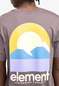 Grijze t-shirt met korte mouwen met een groot grafisch ontwerp van een zon die opkomt boven bergen in geel, blauw en paars met de tekst "element" eronder.