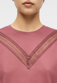 Κοντινό πλάνο ενός ατόμου που φοράει ροζ τοπ με λεπτομέρεια από δαντέλα σε σχήμα V στο στήθος και στρογγυλή λαιμόκοψη.