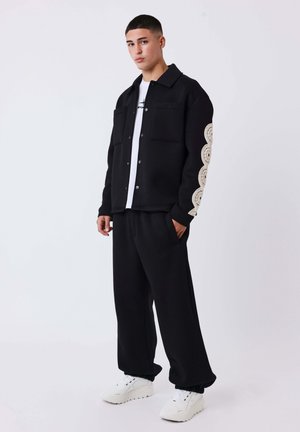 Zwarte fleece jas met twee borstzakken, gedetailleerd crèmekleurig golfpatroon op de mouwen, gecombineerd met zwarte broek en witte sneakers.