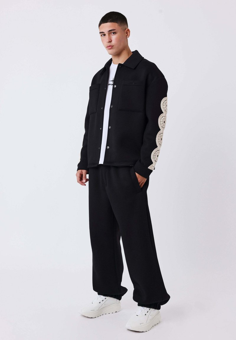 Zwarte fleece jas met twee borstzakken, gedetailleerd crèmekleurig golfpatroon op de mouwen, gecombineerd met zwarte broek en witte sneakers.