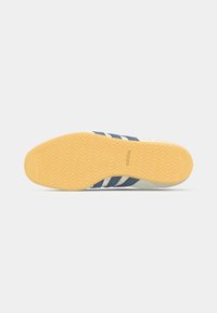 Pohja Adidas lenkkarista, jossa on kuminen ura, tummansiniset raidat ja vaaleanbeige päällinen, nähtynä alhaalta valkoista taustaa vasten.