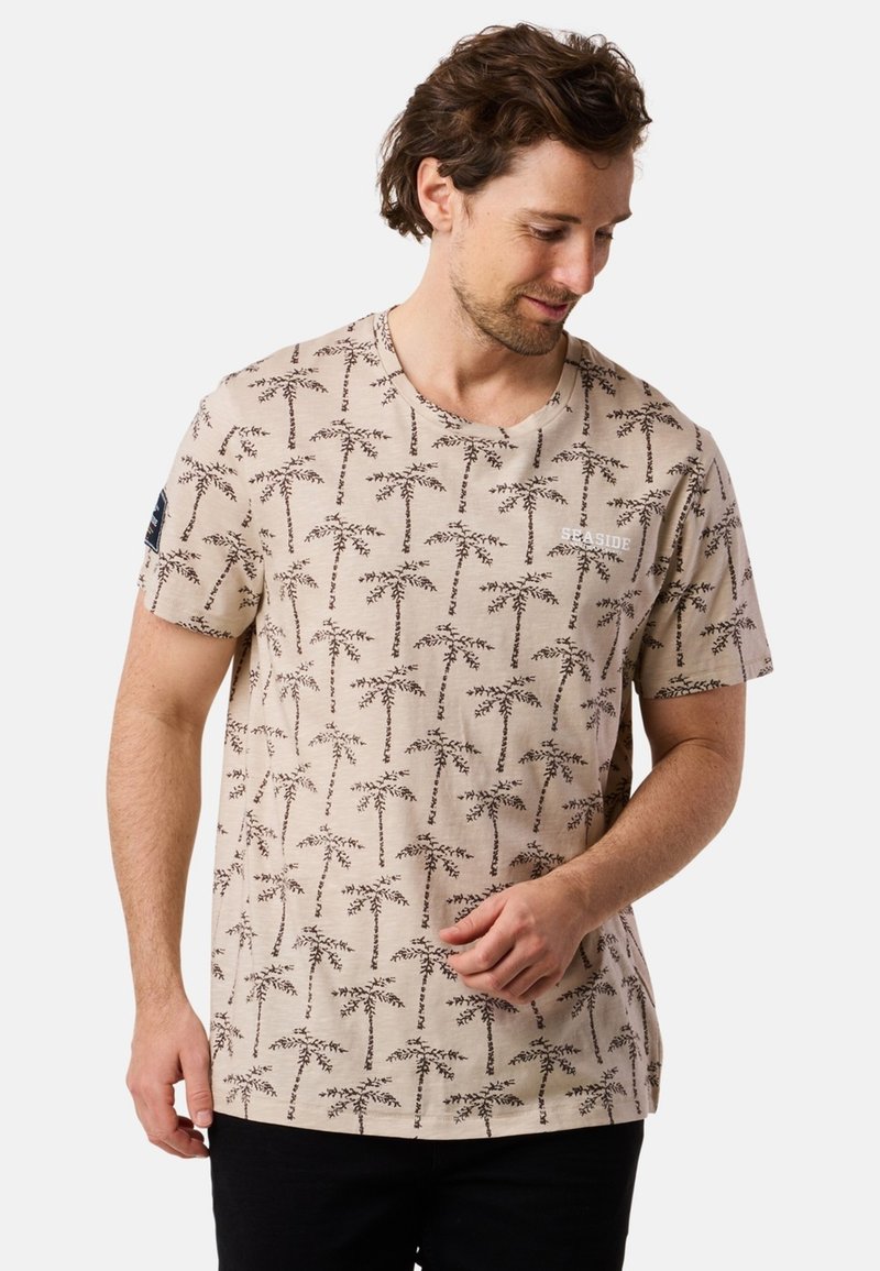 Beige t-shirt featuring a repeated palm tree print in brown, short sleeves, and a crew neckline. Includes a small "SEASIDE" logo on the front.