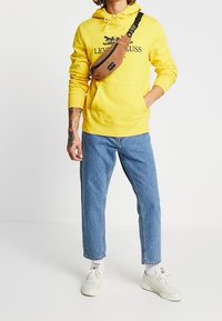 Sudadera amarilla con bolsillo de canguro, logo marino; jeans ajustados de color azul claro; bolso cruzado marrón; zapatillas blancas con acentos grises.