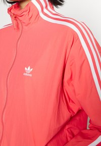 Kvinna som bär en ljus korallfärgad zip-up Adidas träjacka med vita ränder på axlarna och ärmarna.