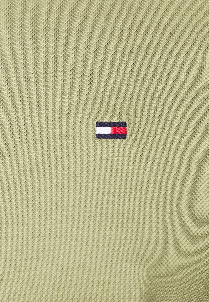 Petit logo rectangulaire rouge, blanc et marine brodé sur un fond de tissu tricoté texturé vert clair.
