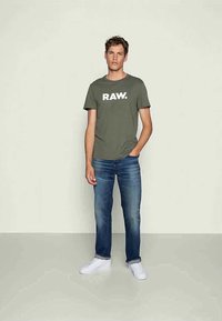 T-shirt verde-oliva com o logótipo branco "RAW.", combinada com jeans azuis claros e ténis brancos; ajuste casual, mangas curtas e design simples.