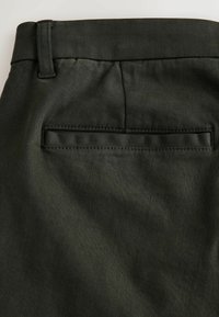 Dark green pants with a textured fabric, featuring a back pocket with a horizontal opening and stitched detailing along the edges.