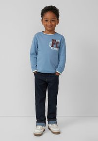Hellblaues Langarmshirt mit "FC DINO KICKERS" Grafik; dunkle Denim-Jeans; weiße Sneakers mit grauen Akzenten; entspannte Passform.