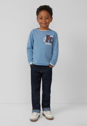 Hellblaues Langarmshirt mit "FC DINO KICKERS" Grafik; dunkle Denim-Jeans; weiße Sneakers mit grauen Akzenten; entspannte Passform.