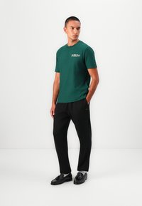 Grøn bomulds t-shirt med korte ærmer, rund halsudskæring og et lille logo trykt på brystet, parret med sorte bukser og sorte sko.