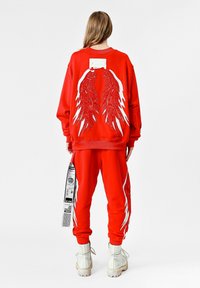 Conjunto de sudadera roja y pantalones, con diseños de alas blancas en la parte posterior y en las piernas exteriores, etiqueta de tela adjunta a los pantalones, usado con botas blancas.