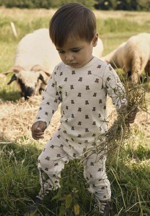 Bela, dolga majica z dolgimi rokavi z motivom medvedka, ki jo nosi otrok na travnati površini. Otrok drži suho travo v eni roki.