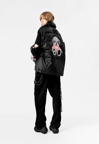 Chaleco de plumas negro con un parche de oso de peluche en rosa y gris en la espalda, llevado sobre una camiseta negra. Combinado con pantalones negros y zapatos gruesos.