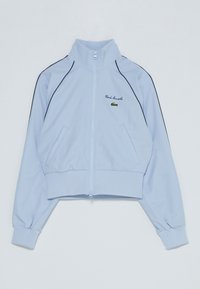 Chaqueta corta de color azul claro, hecha de tela suave, con un alto cuello, cierre de cremallera, puños elásticos y ribetes laterales negros. Logo de Lacoste bordado.