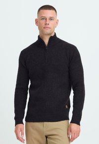 Schwarzer gerippter Strickpullover mit hohem Kragen und Reißverschluss. Seitliches Logodetail. Enganliegende Ärmel und Saum. Getragen über hellbeigen Hosen.