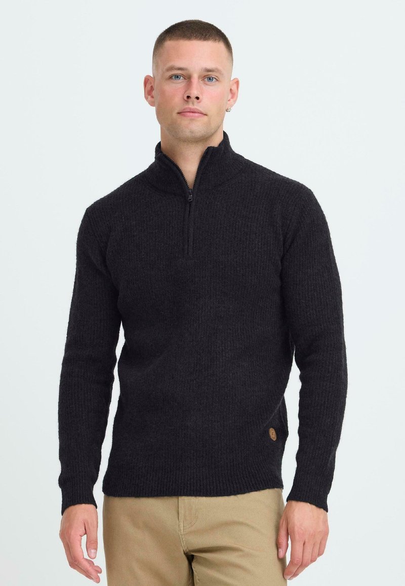 Schwarzer gerippter Strickpullover mit hohem Kragen und Reißverschluss. Seitliches Logodetail. Enganliegende Ärmel und Saum. Getragen über hellbeigen Hosen.