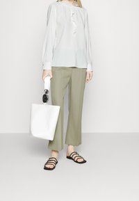 Blouse blanche à volants, pantalon large vert clair, sandales noires à fines lanières, et sac fourre-tout blanc angulaire avec une poignée circulaire.