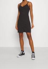 Black ribbed dress with a button-down front, thin straps, and a fitted shape. Worn with black high-top sneakers.