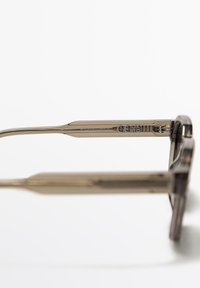 Occhiali da vista di colore marrone, con una spessa montatura rettangolare, aste in metallo e dettagli incisi sulla parte laterale. Texture liscia, design minimalista.