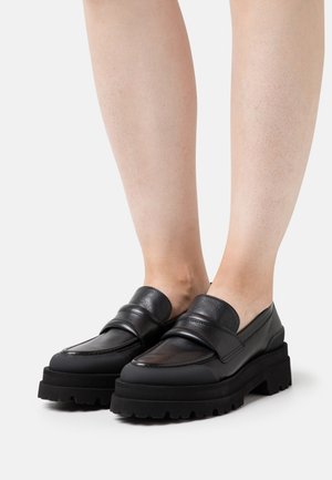 Mustat nahkaiset loaferit, joissa on paksu, kuvioitu musta korokepohja. Kengissä on pyöreä kärki ja näkyvä, leveä remmi päkiän yli.