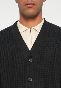 Zwarte gestructureerde cardigan met een diepe V-hals, voorzien van drie zwarte knopen en een crèmekleurig overhemd met hoge kraag eronder.