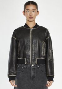 Giacca bomber in pelle nera con design crop, cuciture in contrasto, colletto a coste e tasche laterali con zip. Indossata su jeans scuri.