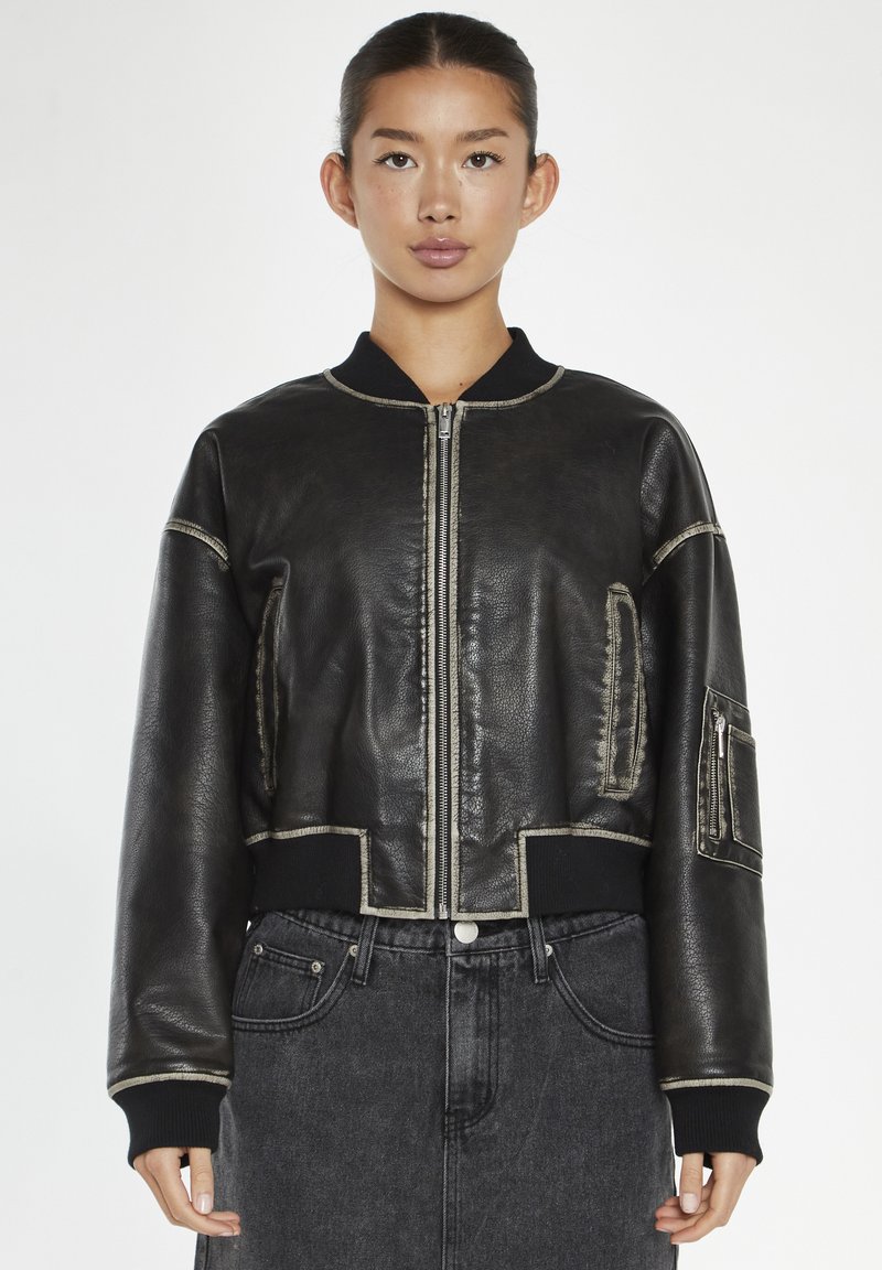 Giacca bomber in pelle nera con design crop, cuciture in contrasto, colletto a coste e tasche laterali con zip. Indossata su jeans scuri.