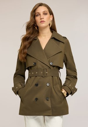 Kobieta w oliwkowozielonym trenczu z paskiem, czarnymi guzikami, złotą bransoletką i kolczykami, pozująca z rękami w kieszeniach.