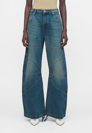 Jean bleu à jambes larges avec des panneaux cousus, porté avec des chaussures blanches pointues et un haut beige.