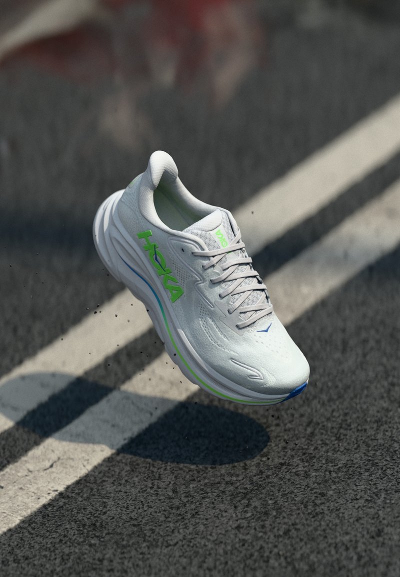 Chaussure de course blanche avec une surface texturée, des accents verts et bleus, une semelle surélevée et une tige en mesh respirant. Présente un logo de marque bien visible.
