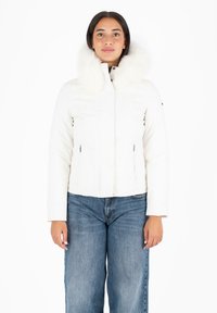 Chaqueta de plumas blanca con cuello de pelo, con un diseño ajustado, bolsillos con cremallera y una longitud corta. Se lleva con jeans de pierna ancha azules.