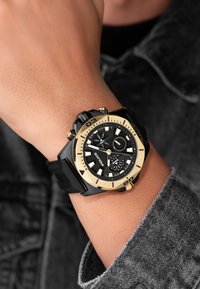Orologio cronografo nero e oro con quadrante nero testurizzato, indicatori delle ore prominenti e bracciale in silicone nero, mostrato su un polso.