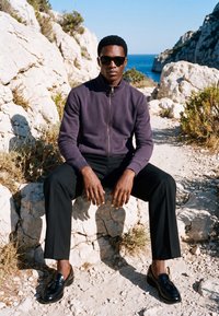 Homme portant des lunettes de soleil, une veste sombre et un pantalon noir assis sur un rocher dans une zone côtière rocheuse avec la mer bleue en arrière-plan.