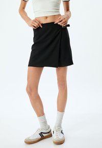 Zwarte wikkelrok met een accent aan de zijkant van een gesp. Heeft een op maat gemaakte stijl en een zachte textuur, gecombineerd met witte sokken en sneakers.