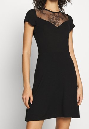 Femme portant une courte robe noire avec des manches courtes et un col en dentelle transparente, debout avec une main détendue le long du corps.