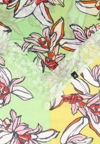 Tissu à motif floral avec de grandes fleurs roses, blanches et rouges sur des fonds verts et jaunes, avec une petite étiquette de marque noire.