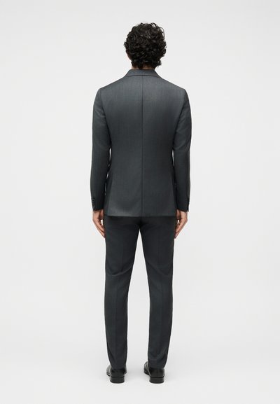 Άνδρας που στέκεται με την πλάτη, φορώντας ένα σκούρο γκρι κοστούμι με ταιριαστό παντελόνι και μαύρα επίσημα παπούτσια σε απλό λευκό φόντο.
