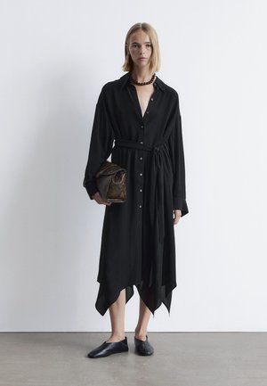 Schwarzes Hemdkleid mit langen Ärmeln und Bindegürtel, mit asymmetrischem Saum. Kombiniert mit schwarzen Slippern und einer strukturierten braunen Clutch.
