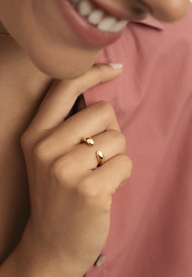 Anelli d'oro con forme morbide e curve indossati sulle punte delle dita, su uno sfondo di tessuto rosa tenue. Sono visibili la texture della pelle e la lucentezza del metallo lucido.