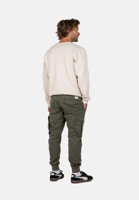 Sudadera beige, pantalones cargo verde oliva con bolsillos con cremallera, puños ajustados y zapatillas negras. Vista desde atrás.