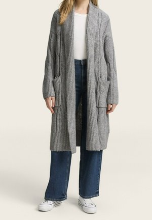Femme portant un long cardigan gris en maille avec des poches, un haut blanc, un jean bleu à jambes larges et des baskets blanches sur un fond neutre.