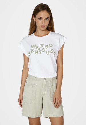Junge Frau trägt ein weißes ärmelloses Oberteil mit der Aufschrift "WHY SO SERIOUS?" und hellbeige hoch taillierte Shorts, steht vor einem schlichten Hintergrund.