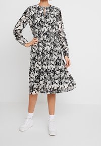 Blommönstrad svartvit klänning med långa ärmar, samlad midja, nyckelhålsringning och en flödande kjol. I kombination med vita sneakers.