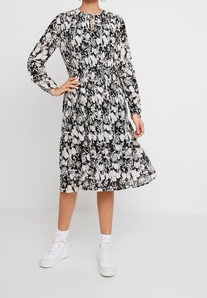 Robe à motif floral noir et blanc avec des manches longues, taille froncée, décolleté en trou de serrure et jupe fluide. Associée à des baskets blanches.