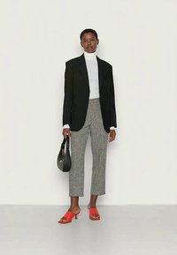 Zwarte oversized blazer, witte coltrui, gestructureerde geruite broek, rode open teen hakken en zwarte handtas. Minimalistisch ontwerp met scherpe lijnen.