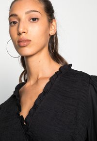 Blusa preta com corpo texturizado e decote franzido. Modelo usou grandes brincos de argola prateados. Fundo neutro.
